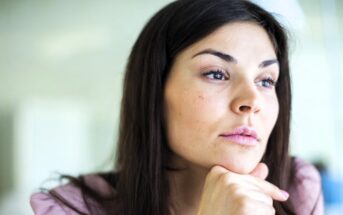 A woman with long dark hair resting her chin on her hand looks thoughtfully into the distance. She has a calm expression, wearing a light pink top, with a soft-focus background.