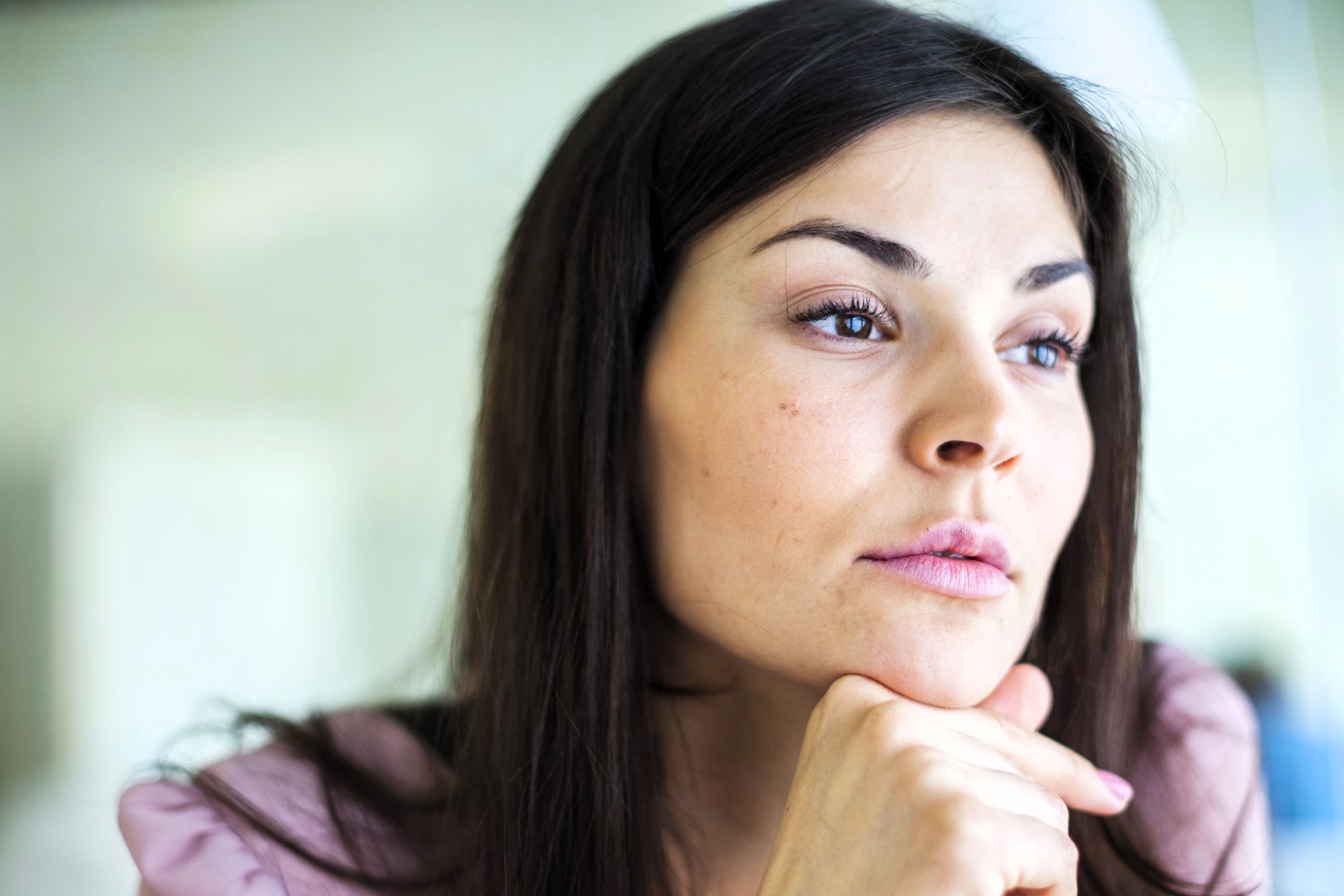 A woman with long dark hair resting her chin on her hand looks thoughtfully into the distance. She has a calm expression, wearing a light pink top, with a soft-focus background.