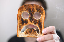 A person holds up a slice of burnt toast with a face-like pattern of holes and dark patches, resembling a sad expression. The background is blurred, and the person's face is not visible. Smoke can be seen drifting from the toast.