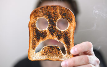 A person holds up a slice of burnt toast with a face-like pattern of holes and dark patches, resembling a sad expression. The background is blurred, and the person's face is not visible. Smoke can be seen drifting from the toast.