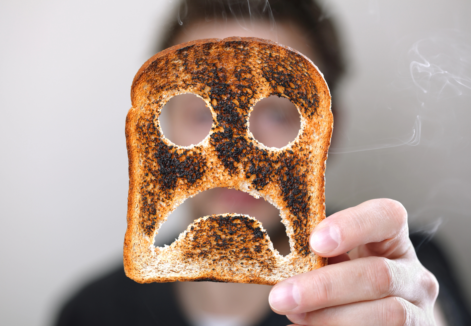 A person holds up a slice of burnt toast with a face-like pattern of holes and dark patches, resembling a sad expression. The background is blurred, and the person's face is not visible. Smoke can be seen drifting from the toast.