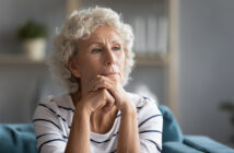 An elderly woman with short curly white hair is seated and gazing thoughtfully into the distance. She rests her chin on her hands, which are clasped together. She is wearing a striped white and navy blue shirt. The background is softly blurred, suggesting an indoor setting.