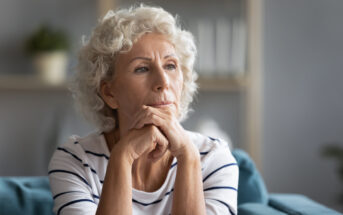 An elderly woman with short curly white hair is seated and gazing thoughtfully into the distance. She rests her chin on her hands, which are clasped together. She is wearing a striped white and navy blue shirt. The background is softly blurred, suggesting an indoor setting.