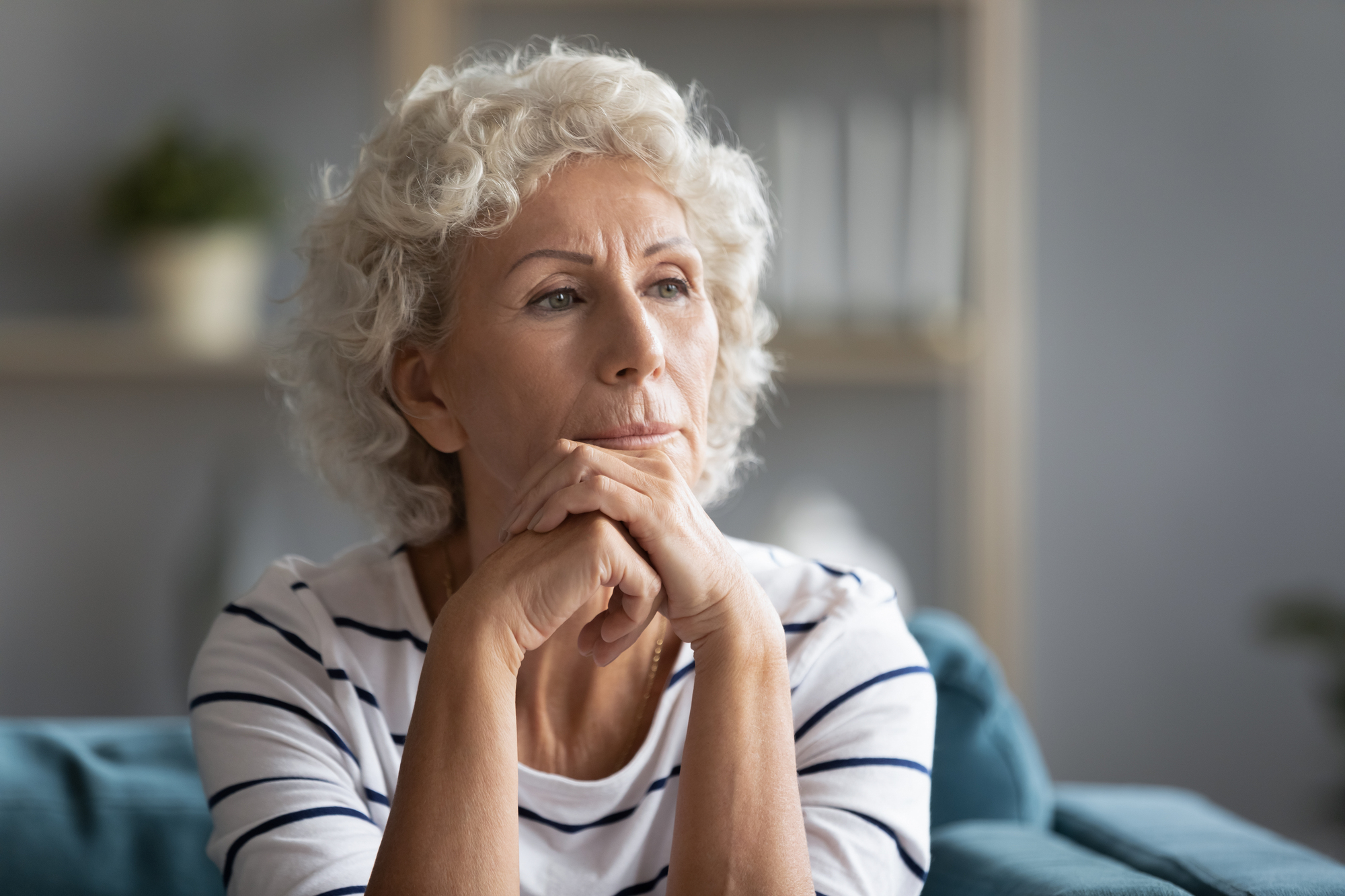 An elderly woman with short curly white hair is seated and gazing thoughtfully into the distance. She rests her chin on her hands, which are clasped together. She is wearing a striped white and navy blue shirt. The background is softly blurred, suggesting an indoor setting.