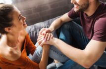 A smiling man and woman sit closely, holding hands and looking at each other affectionately. The woman is wearing an orange off-shoulder top and jeans, while the man is in a maroon t-shirt and jeans. They are sitting on a cozy couch in a warmly lit room.