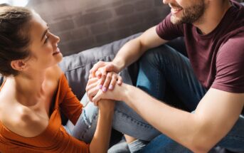 A smiling man and woman sit closely, holding hands and looking at each other affectionately. The woman is wearing an orange off-shoulder top and jeans, while the man is in a maroon t-shirt and jeans. They are sitting on a cozy couch in a warmly lit room.