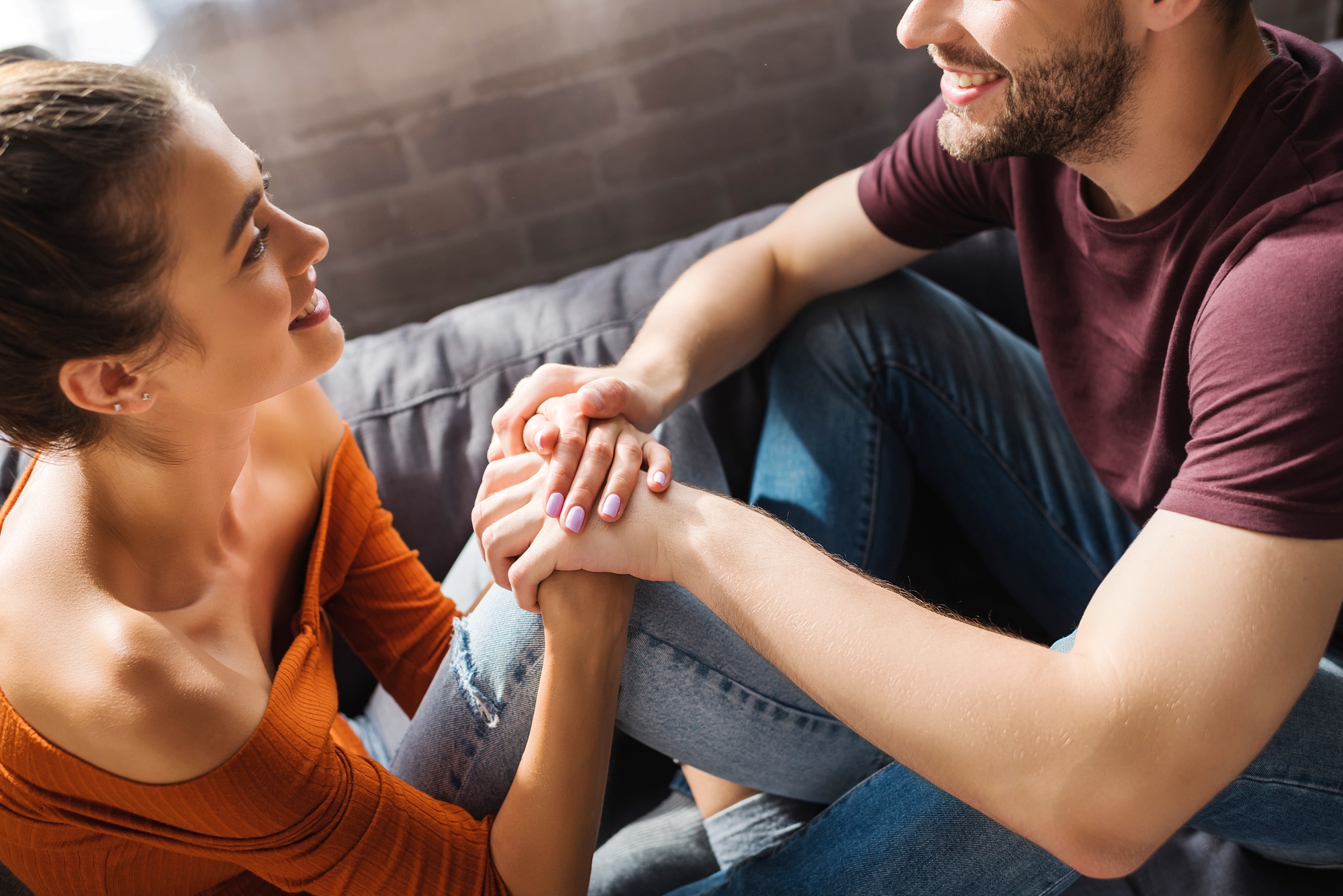 A smiling man and woman sit closely, holding hands and looking at each other affectionately. The woman is wearing an orange off-shoulder top and jeans, while the man is in a maroon t-shirt and jeans. They are sitting on a cozy couch in a warmly lit room.