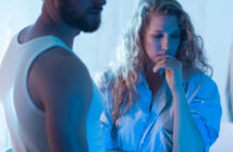 A woman with wavy blonde hair, wearing a blue shirt, looks down pensively while resting her chin on her hand. A man, with a beard and wearing a white tank top, stands partially visible beside her. The scene is lit with a soft blue hue, creating a somber mood.