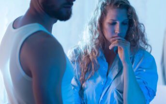 A woman with wavy blonde hair, wearing a blue shirt, looks down pensively while resting her chin on her hand. A man, with a beard and wearing a white tank top, stands partially visible beside her. The scene is lit with a soft blue hue, creating a somber mood.