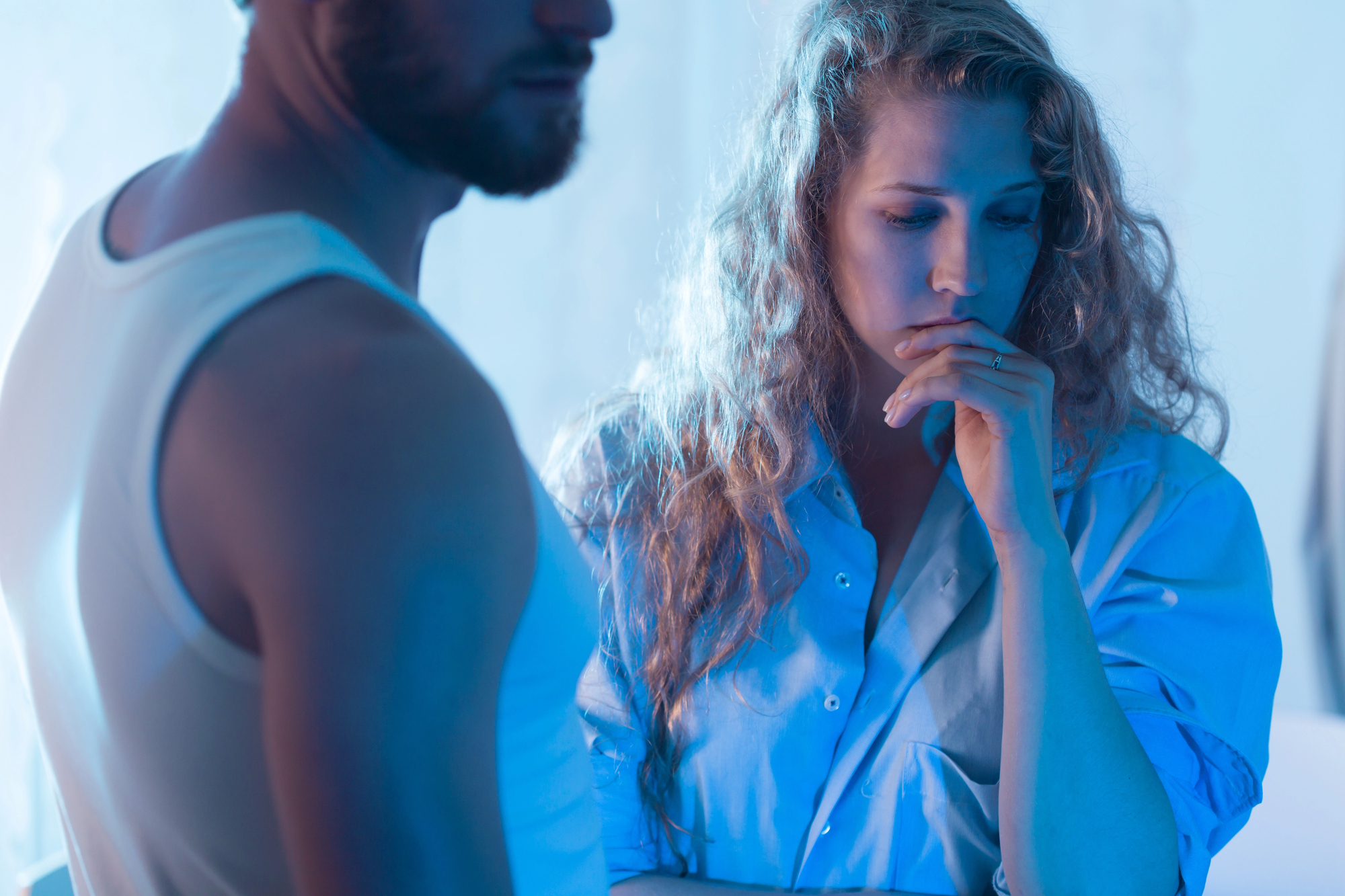 A woman with wavy blonde hair, wearing a blue shirt, looks down pensively while resting her chin on her hand. A man, with a beard and wearing a white tank top, stands partially visible beside her. The scene is lit with a soft blue hue, creating a somber mood.