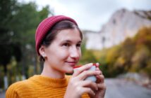 A person wearing a red beanie and a mustard-colored sweater is holding a cup close to their face while smiling slightly. The background features an outdoor scene with trees and a rocky hillside.