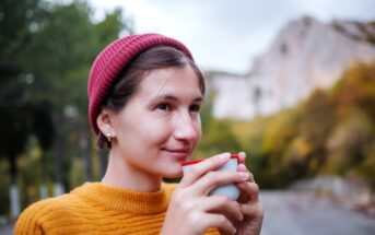 A person wearing a red beanie and a mustard-colored sweater is holding a cup close to their face while smiling slightly. The background features an outdoor scene with trees and a rocky hillside.