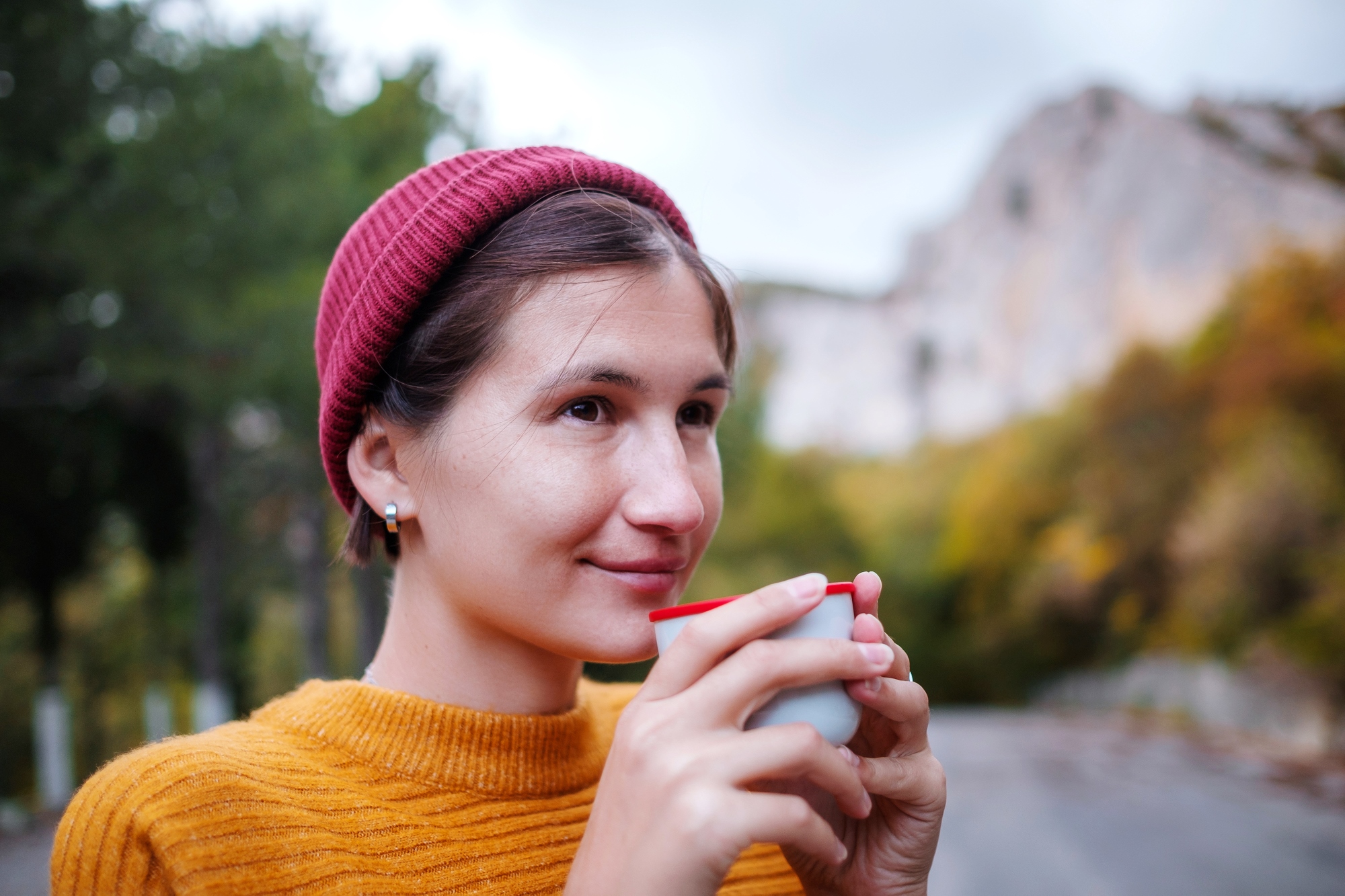 A person wearing a red beanie and a mustard-colored sweater is holding a cup close to their face while smiling slightly. The background features an outdoor scene with trees and a rocky hillside.