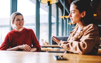 Two women are seated at a wooden table in a modern café with large windows. The woman on the left is wearing a red sweater and smiling, while the woman on the right, in a brown jacket, is gesturing with her hands as she speaks.