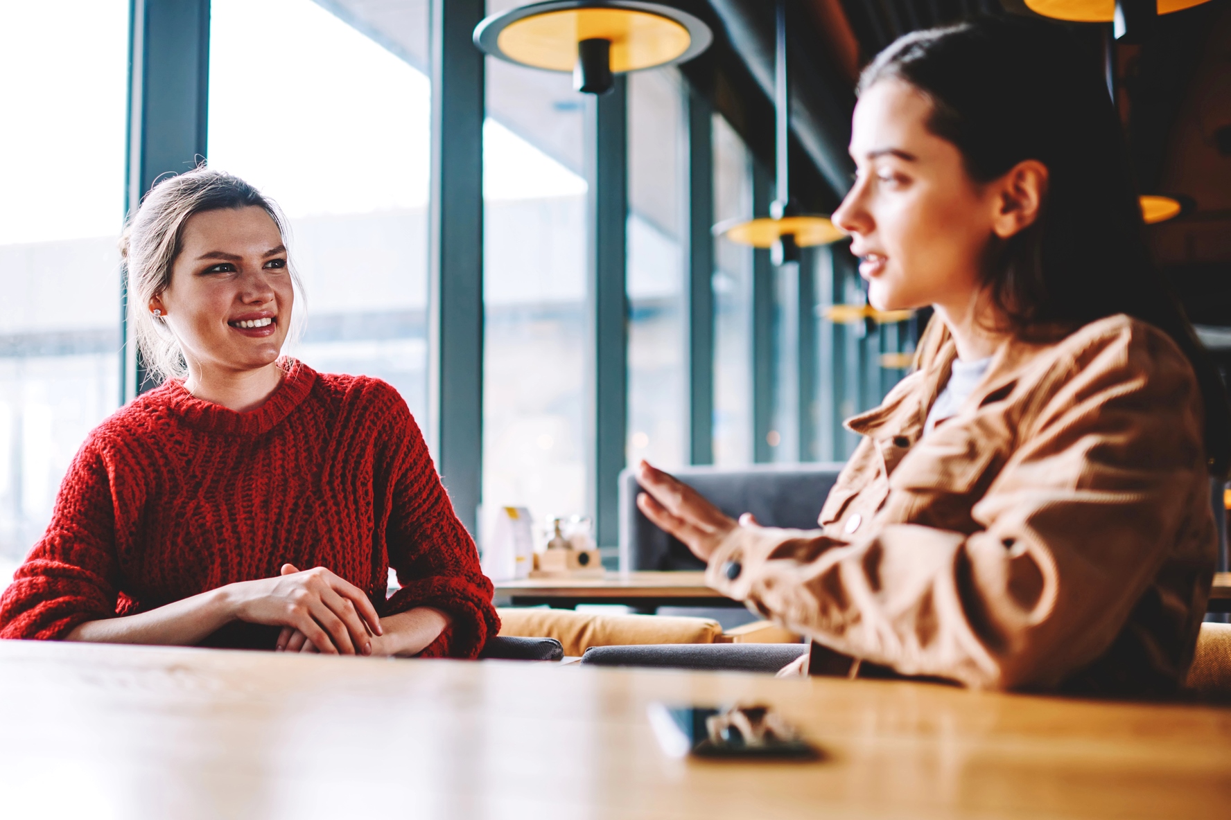 Two women are seated at a wooden table in a modern café with large windows. The woman on the left is wearing a red sweater and smiling, while the woman on the right, in a brown jacket, is gesturing with her hands as she speaks.