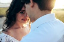 A woman with wavy dark hair gazes at the camera with a gentle smile as a man in a white shirt, whose face is partially obscured, stands close to her. They are outdoors in a sunlit setting, creating a warm, intimate atmosphere.