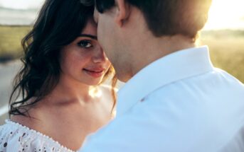 A woman with wavy dark hair gazes at the camera with a gentle smile as a man in a white shirt, whose face is partially obscured, stands close to her. They are outdoors in a sunlit setting, creating a warm, intimate atmosphere.