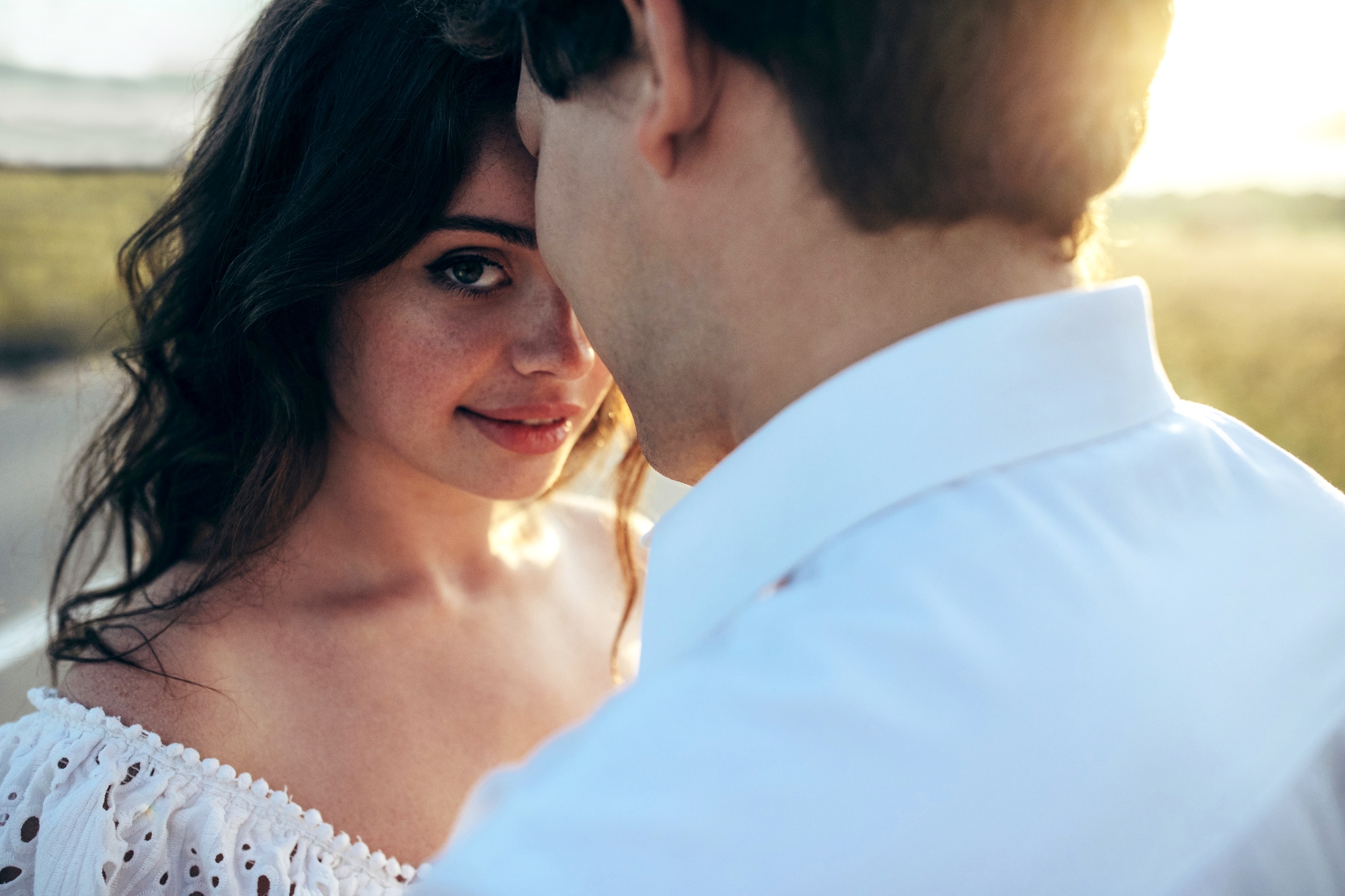 A woman with wavy dark hair gazes at the camera with a gentle smile as a man in a white shirt, whose face is partially obscured, stands close to her. They are outdoors in a sunlit setting, creating a warm, intimate atmosphere.