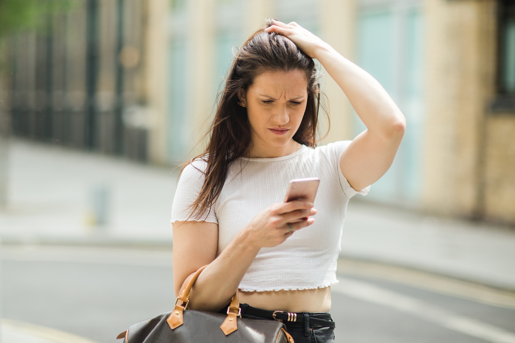 A young woman with long hair, wearing a white crop top and holding a brown handbag, stands on a city street looking at her phone with a concerned expression while touching her head with her other hand.