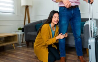 A woman in a mustard sweater kneels on the floor, crying and clinging to the leg of a man standing beside her with a suitcase. The scene appears to take place in a living room with a couch and lamp in the background.