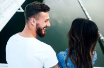 A man with a beard in a white t-shirt and a woman with long brown hair in a denim jacket are seated close together near the edge of a calm body of water, smiling and looking at each other. The background includes parts of a white structure and the reflection in the water.