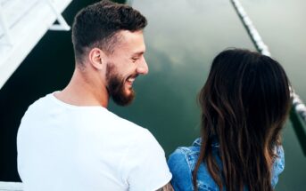 A man with a beard in a white t-shirt and a woman with long brown hair in a denim jacket are seated close together near the edge of a calm body of water, smiling and looking at each other. The background includes parts of a white structure and the reflection in the water.