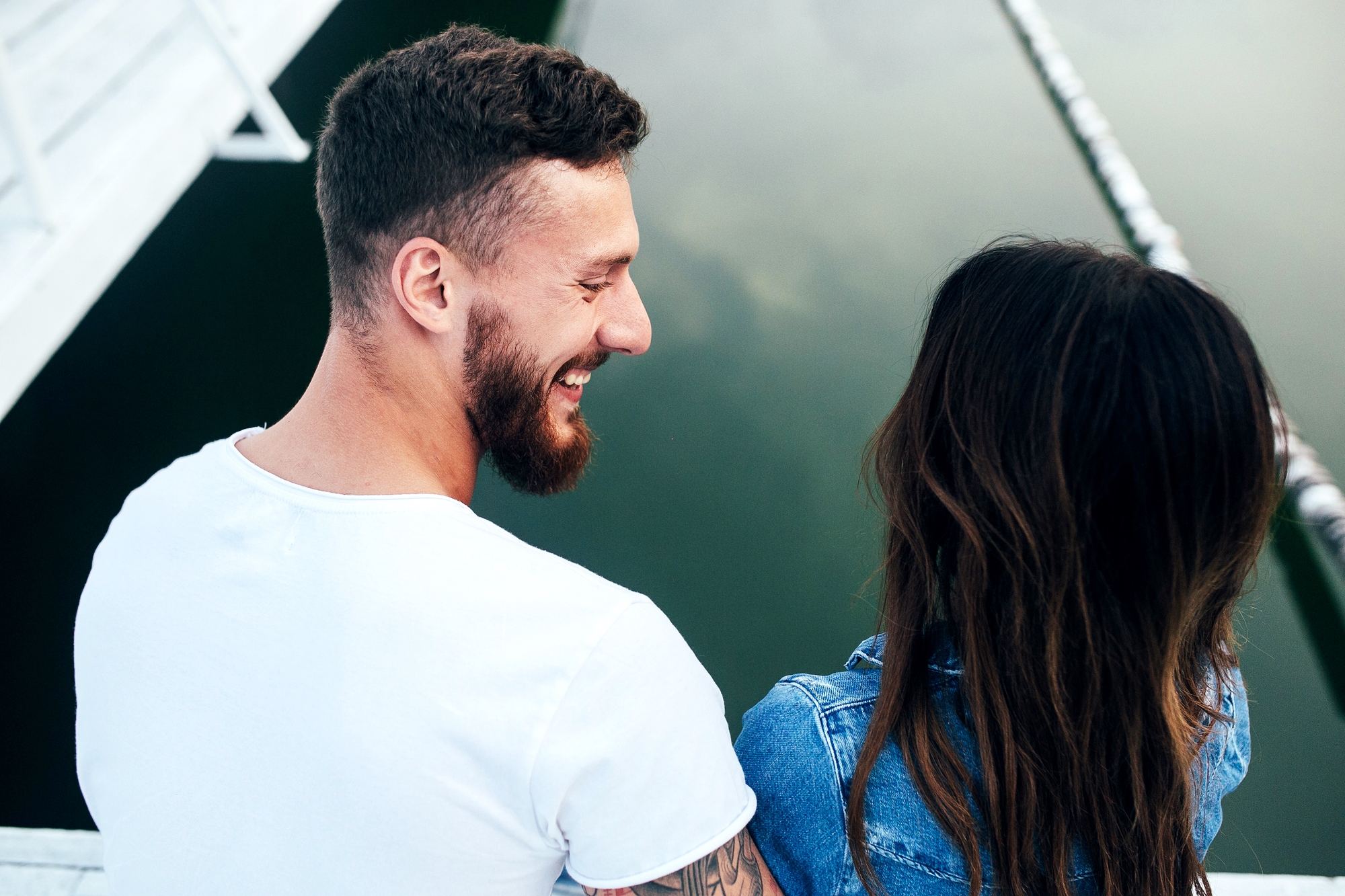 A man with a beard in a white t-shirt and a woman with long brown hair in a denim jacket are seated close together near the edge of a calm body of water, smiling and looking at each other. The background includes parts of a white structure and the reflection in the water.