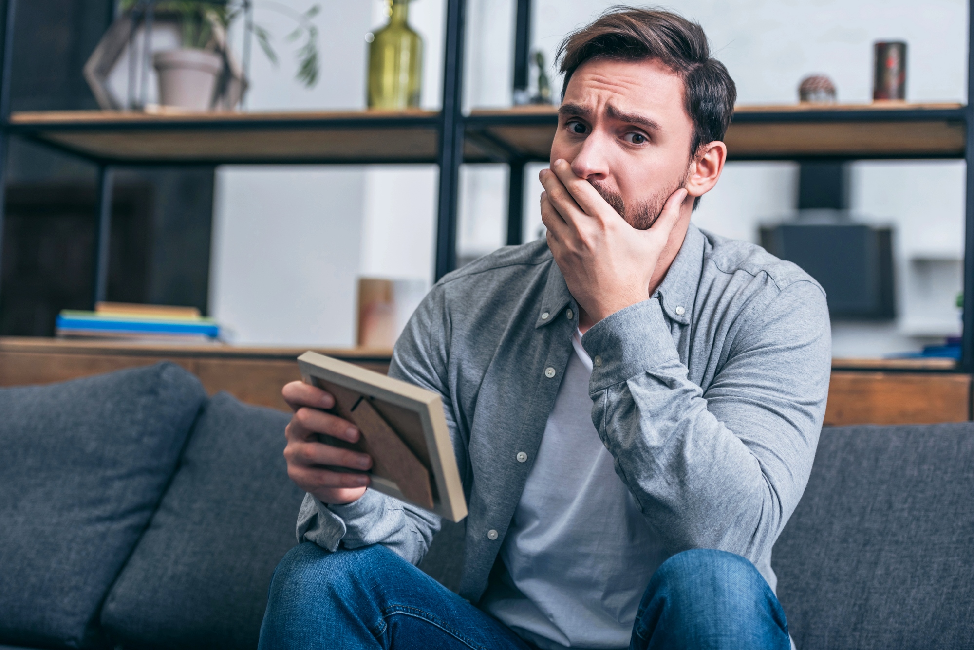 A man sits on a sofa, holding a framed photo with a thoughtful and concerned expression. His left hand partially covers his mouth as he looks into the distance. The background shows shelves with decorative items and books.