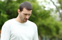 A man with short dark hair and a beard looks down with a serious expression. He is wearing a light gray, long-sleeve shirt and standing outdoors. The background is blurred, showing lush greenery.