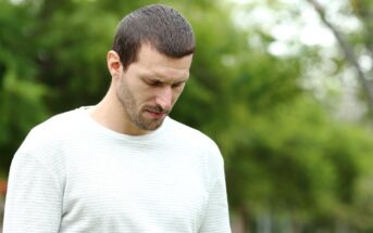 A man with short dark hair and a beard looks down with a serious expression. He is wearing a light gray, long-sleeve shirt and standing outdoors. The background is blurred, showing lush greenery.