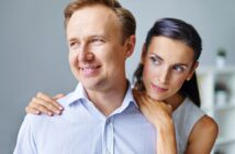 A man wearing a light blue shirt smiles while looking off to the side, with a woman standing behind him, gently embracing him around the shoulders. The woman has dark hair and appears to be gazing at something in the distance. They are indoors with a neutral background.