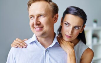 A man wearing a light blue shirt smiles while looking off to the side, with a woman standing behind him, gently embracing him around the shoulders. The woman has dark hair and appears to be gazing at something in the distance. They are indoors with a neutral background.