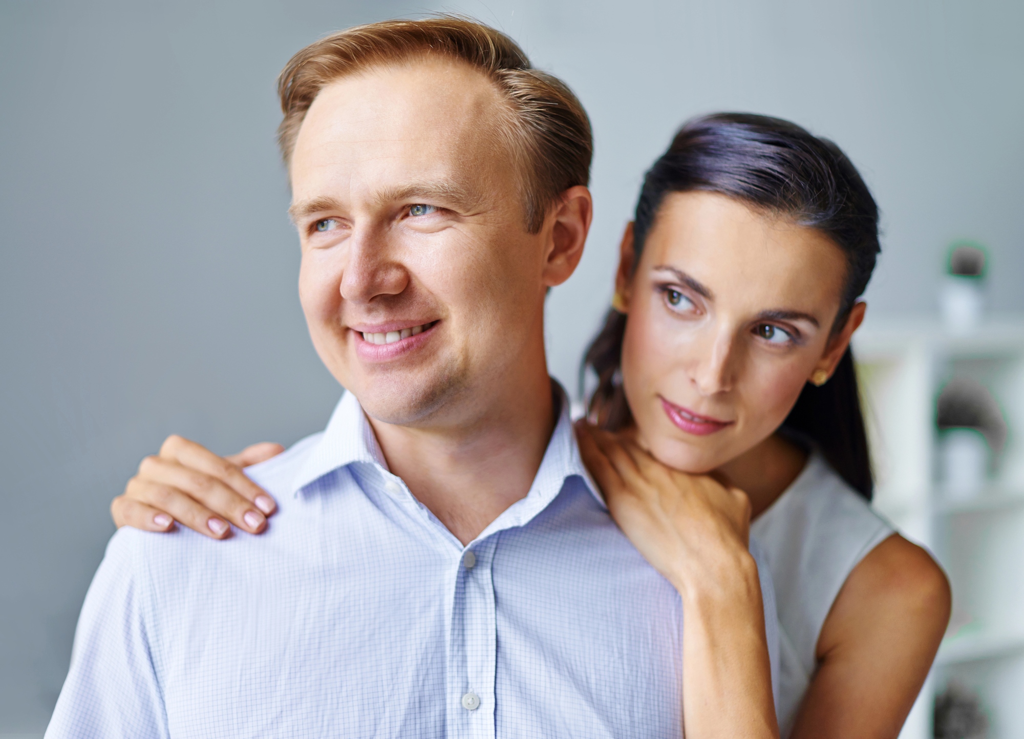 A man wearing a light blue shirt smiles while looking off to the side, with a woman standing behind him, gently embracing him around the shoulders. The woman has dark hair and appears to be gazing at something in the distance. They are indoors with a neutral background.