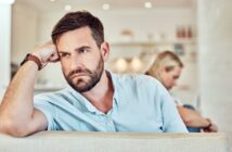 A man with a beard and short hair rests his head on his hand, looking pensive while seated on a couch. In the blurred background, a woman with blonde hair sits turned away from him, also lost in thought. The setting appears to be a modern living room.