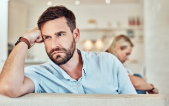 A man with a beard and short hair rests his head on his hand, looking pensive while seated on a couch. In the blurred background, a woman with blonde hair sits turned away from him, also lost in thought. The setting appears to be a modern living room.