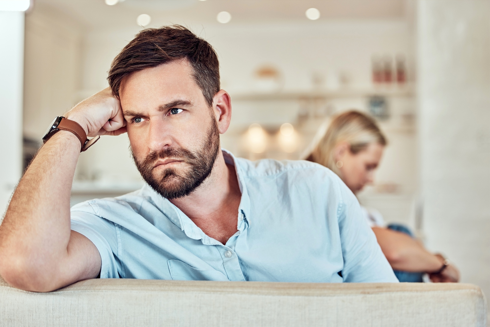 A man with a beard and short hair rests his head on his hand, looking pensive while seated on a couch. In the blurred background, a woman with blonde hair sits turned away from him, also lost in thought. The setting appears to be a modern living room.