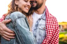 A couple embraces outdoors, smiling warmly. The woman is wearing a denim jacket, and the man has a light denim shirt layered under a red and white checkered blanket. The background is softly blurred, suggesting a peaceful and sunny rural setting.