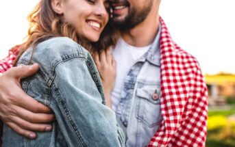 A couple embraces outdoors, smiling warmly. The woman is wearing a denim jacket, and the man has a light denim shirt layered under a red and white checkered blanket. The background is softly blurred, suggesting a peaceful and sunny rural setting.