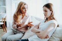 A woman with blonde hair, wearing a pink blouse, is sitting on a couch gesturing as she speaks. Next to her, a younger woman with brown hair in a white t-shirt is sitting with arms crossed, looking down and away. They both appear deep in conversation.