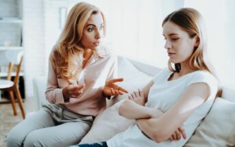 A woman with blonde hair, wearing a pink blouse, is sitting on a couch gesturing as she speaks. Next to her, a younger woman with brown hair in a white t-shirt is sitting with arms crossed, looking down and away. They both appear deep in conversation.