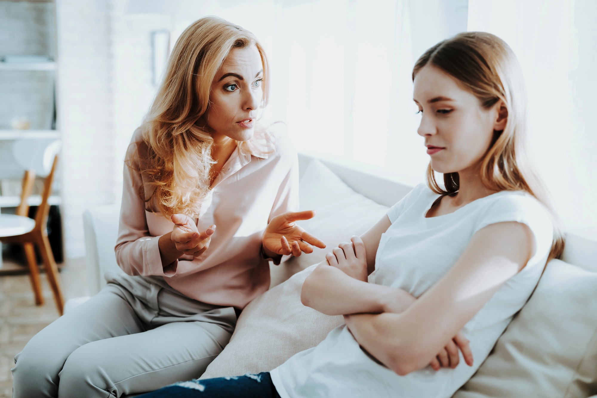 A woman with blonde hair, wearing a pink blouse, is sitting on a couch gesturing as she speaks. Next to her, a younger woman with brown hair in a white t-shirt is sitting with arms crossed, looking down and away. They both appear deep in conversation.