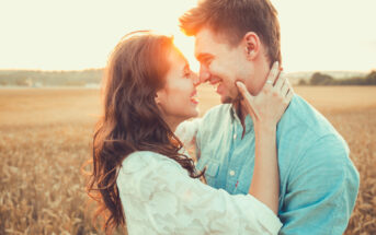 A couple stands in a wheat field at sunset, facing each other closely with their noses touching. The woman is holding the man's face in her hands, both smiling and looking happy. The scene is warm and glowing with the sun in the background.