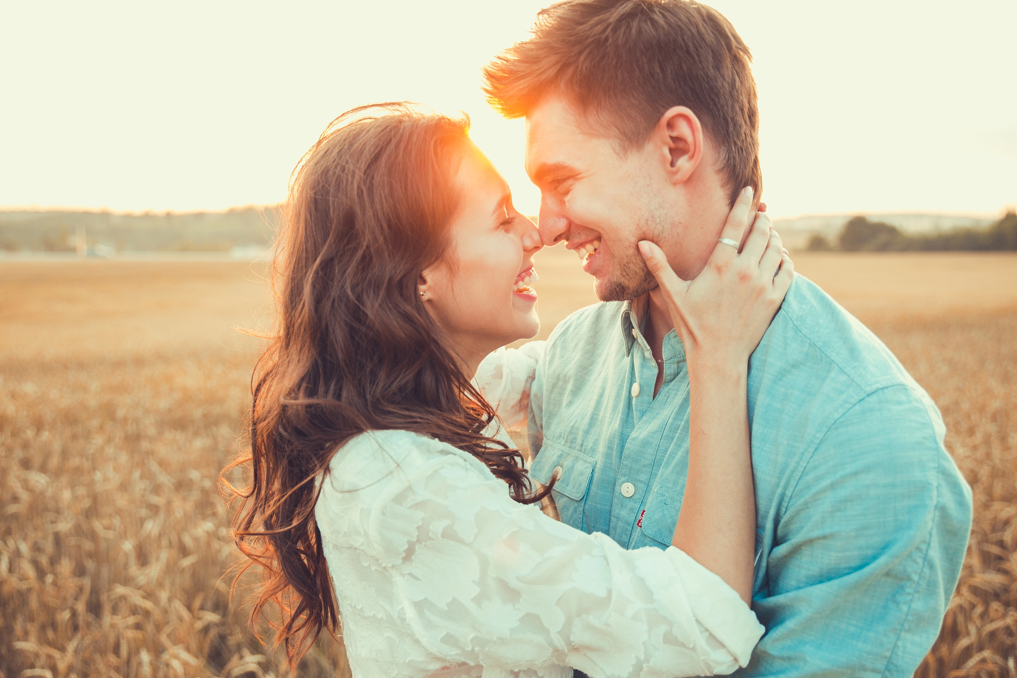 A couple stands in a wheat field at sunset, facing each other closely with their noses touching. The woman is holding the man's face in her hands, both smiling and looking happy. The scene is warm and glowing with the sun in the background.