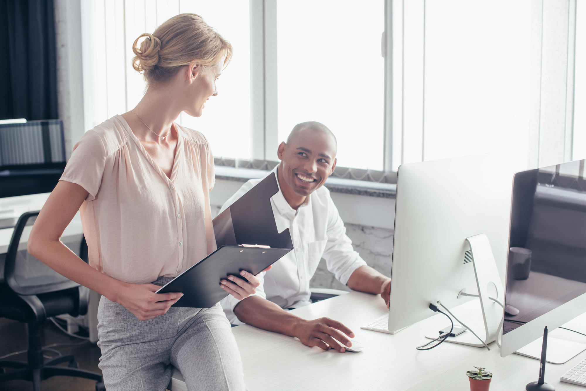 A woman holding a clipboard and wearing business attire is standing and smiling at a man sitting at a desk with a computer. The man is also smiling and looking at the woman. They are in a modern office with large windows. A small potted plant is on the desk.