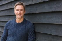 A middle-aged man with short brown hair and a friendly smile leans against a dark wood-paneled wall. He is wearing a blue cable-knit sweater and looking directly into the camera. The lighting is natural, indicating an outdoor setting.