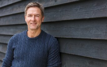 A middle-aged man with short brown hair and a friendly smile leans against a dark wood-paneled wall. He is wearing a blue cable-knit sweater and looking directly into the camera. The lighting is natural, indicating an outdoor setting.