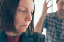 A close-up of a woman with a serious expression, dressed in a blazer, looking downward. In the blurred background, a person in a checkered shirt is seen with one arm raised, suggesting a heated or emotional exchange.