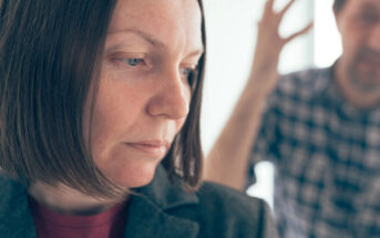 A close-up of a woman with a serious expression, dressed in a blazer, looking downward. In the blurred background, a person in a checkered shirt is seen with one arm raised, suggesting a heated or emotional exchange.