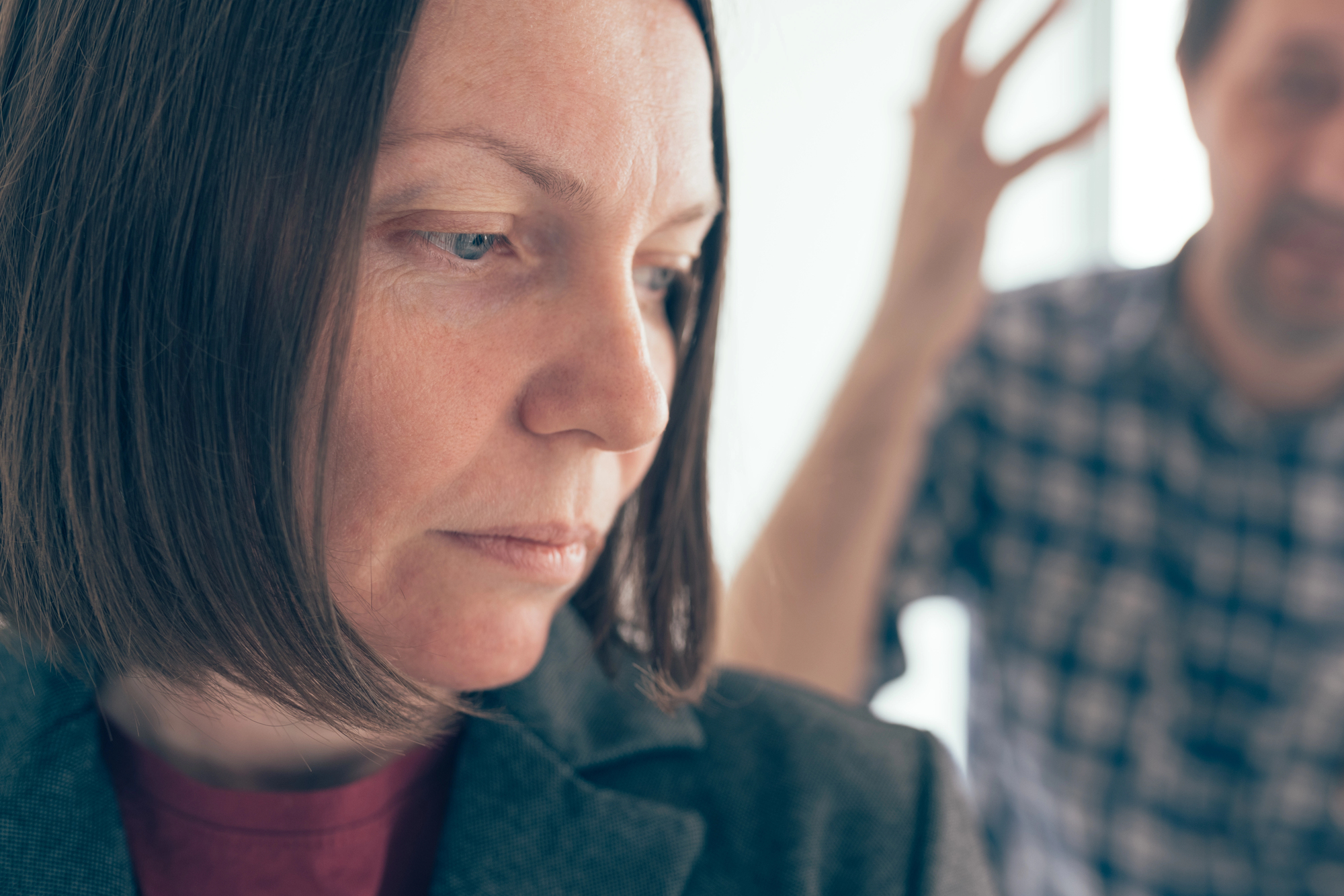 A close-up of a woman with a serious expression, dressed in a blazer, looking downward. In the blurred background, a person in a checkered shirt is seen with one arm raised, suggesting a heated or emotional exchange.