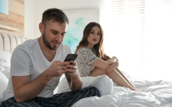 A man in a white t-shirt and plaid pajama pants sits on a bed engrossed in his smartphone. Behind him, a woman in a casual dress sits with her knees drawn up, looking at him with a neutral expression. The room is softly lit by sunlight streaming through the window blinds.